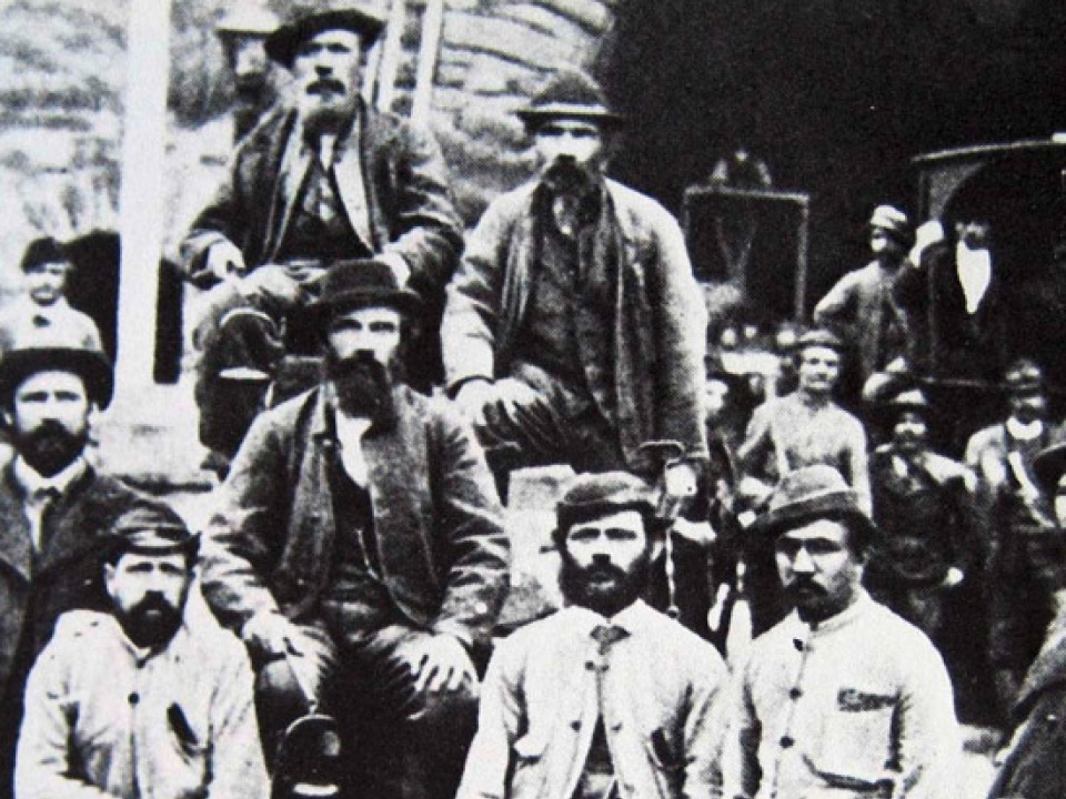 Historisches Schwarz-Weiß-Foto einer Gruppe von Männern mit Bärten, die Hüte und historische Kleidung tragen und im Freien posieren; einige stehen, andere sitzen, mit unscharfen Figuren und Strukturen im Hintergrund.